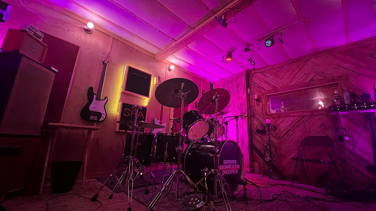 A music rehearsal space at MOON MUTT Recording Studio, featuring a drum set lit by purple stage lights, with guitars hanging on the wall. | MOON MUTT Recording Studio